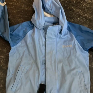 Blå regnkläder  med huva från Exani Stl 104 - Snygg ljusblå regnjacka från Exani med huva och dragkedja framtill. Jackan har två fickor och reflexdetaljer på ärmarna. Perfekt för blöta dagar och enkel att matcha med regnbyxor. Jackan är tillverkad i syntetmaterial och har en normal passform.