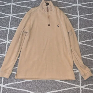 Beige half zip tröja Polo Ralph Lauren - Snygg beige långärmad tröja från Polo Ralph Lauren med half zip och klassisk broderad logga på bröstet. Tröjan har hög krage och är tillverkad i mjuk bomull, perfekt för lager-på-lager under höst och vinter.
