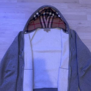 Äkta Burberry kofta - Säljer en grå hoodie från Burberry i storlek M. Tröjan har klassiskt rutigt foder i huvan, snörning och dragkedja framtill. Två fickor på magen och ribbade muddar vid ärmslut och nederkant. Perfekt för dig som gillar stilren design med lyxig touch.