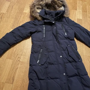 Svart lång dunjacka Rockandblue - Säljer en svart lång dunjacka från Rockandblue med fluffig äkta päls på huvan. Jackan har flera snygga dragkedjor, tryckknappar och quiltad design. Perfekt för kalla dagar med sin varma och stilrena look. Fickor med dragkedja och innerficka för extra funktion.