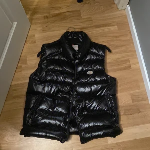 Svart dunväst från Moncler - Snygg svart dunväst från Moncler med glansig finish och klassisk logga på bröstet. Västen har hög krage, dragkedja och knappar framtill samt två fickor med dragkedja. Perfekt för lager-på-lager och streetstyle.