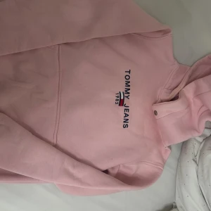 Rosa hoodie från Tommy Jeans - Säljer en ljusrosa hoodie från Tommy Jeans med broderad logga och text på bröstet. Tröjan har huva och klassisk passform, perfekt för en chill och avslappnad stil.