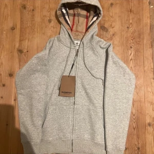 Grå hoodie från Burberry med dragkedja - Snygg grå hoodie från Burberry med klassiskt rutigt foder i huvan och dragkedja framtill. Tröjan har snörning i huvan och ribbade muddar vid ärmslut och nederkant. Perfekt för en avslappnad och stilren look. Sitter riktigt bra på storlek M men sitter som S 