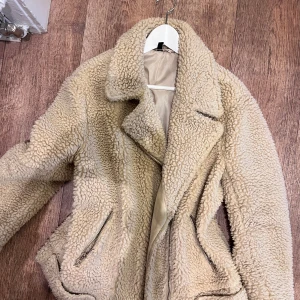 Beige teddyjacka med dragkedjor - Säljer en beige teddyjacka med coola dragkedjor vid fickorna och klassisk krage. Jackan har en fluffig och mysig look, perfekt för att hålla värmen. Materialet är teddy/fleece och modellen är normal i passningen med långa ärmar.
