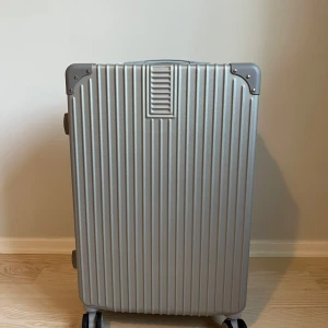 Silver resväska med kodlås - Snygg och stilren resväska i silver med randigt mönster och hårt skal. Väskan har fyra hjul, kodlås och praktiska handtag på både sidan och toppen. Perfekt för dig som vill resa smidigt och ha koll på dina grejer. Riktigt clean look och modern design.