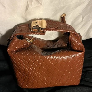 Brun flätad handväska i skinnimitation - Snygg brun handväska med flätad look och guldfärgade detaljer. Väskan har dragkedja upptill och ett brett handtag med spänne. Tillverkad i skinnimitation som ger en lyxig känsla. Perfekt storlek för det viktigaste och passar till många outfits. 12 cm hög💗 17cm bred 💞kommer med avtagbar axelrem 