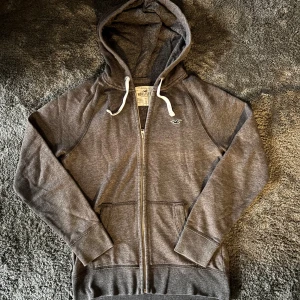 Hollister zip hoodie - Säljer denna schyssta och trendiga gråa zip hoodie från Hollister med vit snörning i huvan. Tröjan har två fickor på magen och diskret Hollister-logga på bröstet. Perfekt för chill dagar.