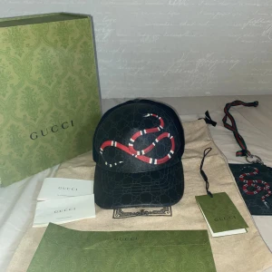 Gucci snake keps - Svart keps från Gucci med klassiskt GG-mönster och en röd, vit och svart orm framtill. Kepsen har mesh på sidorna och justerbart band bak med röd-blå rand. Tillverkad i polyester och bomull, perfekt för en trendig streetwear-look. Kort hållaren ingår i priset kepsen är knappt använd men kort hållaren är endå ganska använd. Hör av dig vid funderingar pris kan diskuteras lite grann 