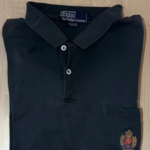 Svart pikétröja från Ralph Lauren - Svart pikétröja från Polo Ralph Lauren med klassisk krage och två knappar framtill. På bröstet finns en ficka med broderat emblem i guld, rött och blått. Tröjan är gjord i mjuk bomull och har korta ärmar, perfekt för en clean och stilren look.