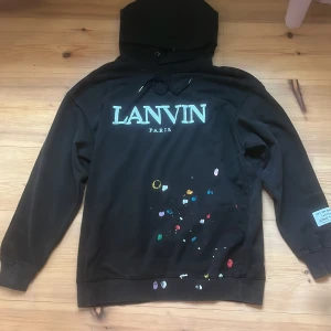 Svart Lanvin x Galerie hoodie - Svart hoodie från Lanvin x DÉPT. de la GALERIE med stor logga framtill och tryck på ryggen. Färgstänk i olika nyanser framtill ger en unik look. Huva med snörning och patch på ärmen med texten 'Art on display courtesy of Josué Thomas'.  Helt ny!