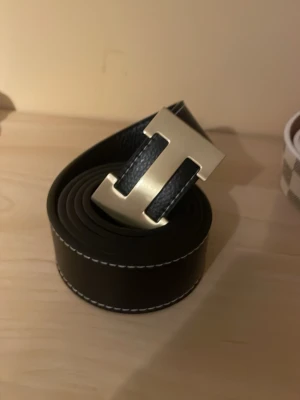 Svart bälte  - Bilden visar ett svart skärp med ett stort H-format spänne i guld, men ingen väst finns med på bilden.