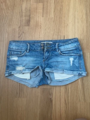 Lågmidjade jeansshorts - Säljer ett par blå jeansshorts med råa kanter och slitna detaljer. Shortsen har dubbla knappar framtill, fem fickor och bälteshällor. Perfekta för sommaren och ger en avslappnad vibe till din outfit.