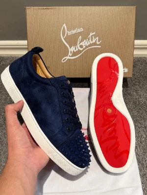 Mörkblå mocka sneakers från Louboutin - Snygga mörkblå sneakers från Christian Louboutin i mocka med vita sulor och den ikoniska röda undersidan. Skorna har snörning och coola nitar på tån för extra edge. Insidan är beige och märkt med Louboutin Paris. Perfekta för dig som vill sticka ut.