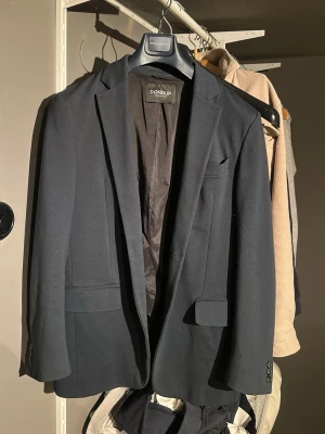 Blå kavaj från Dondup - Säljer en mörkblå blazer från Dondup, tillverkad i Italien. Klassisk modell med smal passform, två fickor med lock och en bröstficka. Kavajen har släta knappar vid ärmsluten och är helfodrad med mjukt tyg. Perfekt för en clean och stilren look. Den är stretchig. 