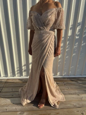 Champagnefärgad långklänning med slits - Elegant offshoulder långklänning i champagnefärg med glittrigt tyg och puffiga korta ärmar. Klänningen har omlottliv, markerad midja och en snygg slits framtill. Perfekt för bal eller festliga tillfällen.