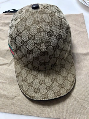 Svart Gucci keps med GG-mönster - Snygg svart keps från Gucci med klassiskt GG-monogram över hela kepsen. Har en grön och röd rand på sidan som ger den typiska Gucci-looken, kommer med tag o dustpåse