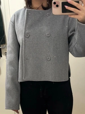 Grå cropped skepparkavaj från Gina Tricot - Säljer en stilren grå skepparkavaj från Gina Tricot. Jackan är croppad med dubbelknäppning och stora knappar framtill. Den har en boxig passform och är gjord i mjukt ullmix-material. Perfekt till lager på lager och ger en clean look.