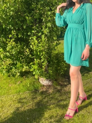 Turkos kortklänning med v-ringning - Säljer en turkos kortklänning med v-ringad hals och långa, transparenta ärmar. Klänningen har markerad midja och ett luftigt, flowy tyg som passar perfekt till varma dagar. Supersnygg och trendig look!