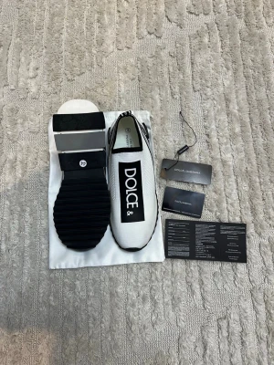 Dolce & Gabbana vita sneakers  - Säljer ett par vita Dolce & Gabbana sneakers med svart logotyptryck på ovansidan och svart-vit detaljerad sula. Skorna har en rund tå och är tillverkade i syntetmaterial med stickad mesh för skön passform. Medföljer originalpåse och certifikat.