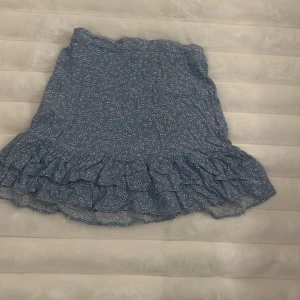 Blommig blå volangkjol från Lindex - Supersöt blå kortkjol från Lindex med små vita blommor och volangkant nertill. Kjolen har resår i midjan för extra bekvämlighet och är perfekt för vår och sommar. Lätt och luftig, passar dig som gillar en feminin och lekfull stil. Ps frakten är med i priset!!!!🩷