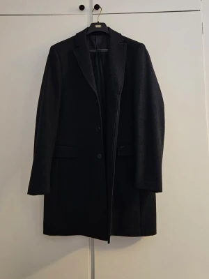 Svart klassisk rock med knappar - Säljer en svart rock från Hampton Republik med klassisk krage och tre knappar framtill. Jackan har två fickor med lock och är helfodrad. Perfekt för dig som vill ha en stilren och tidlös look.