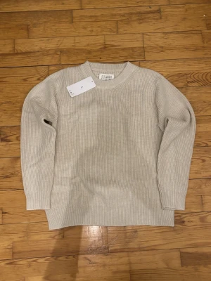 Beige stickad tröja Maison Margiela - Säljer en stilren beige stickad tröja från Maison Margiela. Tröjan har ribbad struktur, rund halsringning och långa ärmar. Perfekt för lager-på-lager och enkel att matcha med allt. Diskret logodetalj i nacken.