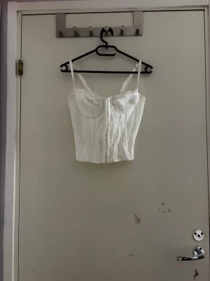 Vit spetskorsett med snörning - Säljer en vit korsett med tunna axelband och vacker spetsdetalj. Framsidan har en stilren design medan baksidan har snygg snörning som ger en justerbar passform. Toppen är croppad och har en feminin v-ringning. Perfekt att styla till jeans eller kjol.