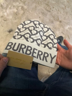 Vit och grå Burberry mössa (äkta) - Säljer en snygg vit och grå mössa från Burberry med ett grafiskt mönster av B-bokstäver och stor logotyp framtill. Mössan är stickad och har en bred uppvikt kant med Burberry-text. Perfekt för dig som gillar lyxiga accessoarer.