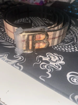 Rutig beige bälte med guldspänne - Säljer ett snyggt bälte med klassiskt rutigt mönster i beige, brunt och svart. Bältet har ett stort guldigt B-spänne framtill och är gjort i ett material som liknar skinn. Perfekt accessoar för att lyfta din outfit.