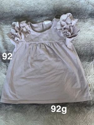 Ljuslila blus med volangärmar - Supersöt ljuslila blus med stora volangdetaljer på axlarna. Blusen har rund halsringning och är kortärmad, perfekt för dig som gillar romantiska detaljer och en söt look.🌸 Fint använt skick. Storlek 92