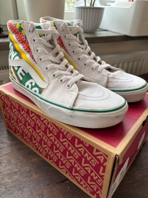 Vita Vans high-top sneakers med färgglada detaljer - Unika Vans sk8-high sneakers i vitt tyg med färgglada detaljer i grönt, gult och rött på sidan och hälen. Skorna har snörning, rund tå och klassisk Vans-sula med grön rand. De är endast använda några gånger och säljes eftersom de är för små nu. Skorna är i storlek 41 och i fint skick. Hör gärna av dig vid frågor.