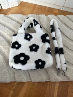 Fluffig blommig axelväska i svartvitt - Supersöt fluffig axelväska i vitt med svarta blommor. Väskan har både handtag och en avtagbar axelrem, samt innerfack med dragkedja. Perfekt för dig som vill ha en unik och mysig accessoar som sticker ut.