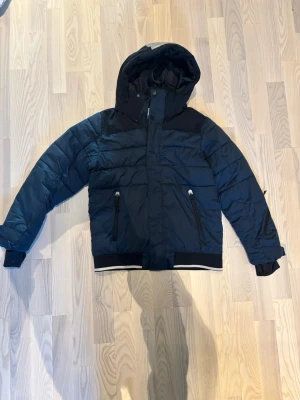 Mörkblå pufferjacka från McKinley - Säljer en mörkblå pufferjacka från McKinley med huva och dragkedja. Jackan har svarta detaljer, två framfickor med dragkedja och en vit rand på huvan samt nederkanten. Perfekt för kalla dagar och riktigt skön passform.