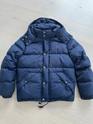 Mörkblå pufferjacka från Polo Ralph Lauren - Snygg mörkblå pufferjacka från Polo Ralph Lauren med huva och dragkedja samt knappar framtill. Insidan har coola gula ränder och en stor 18-tryck. Jackan har två fickor med knappstängning och snörning nedtill. Perfekt för kalla dagar och riktigt sportig vibe.