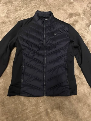 Svart pufferjacka från Calvin Klein - Svart pufferjacka från Calvin Klein Golf med snygga quiltade detaljer på framsidan och diskret CK-logga. Jackan har dragkedja hela vägen, långärmade sidor i mjukt material och hög krage. Perfekt för dig som gillar stilren och sportig look.