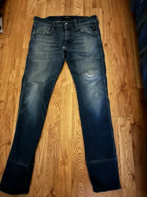Replay jeans blå, storlek 33/34 - Säljer ett par snygga Replay jeans i mörkblå tvätt med slitningar och en liten reva på låret. Klassisk femficksmodell med raka ben och tydliga kontrastsömmar. Jeansen är i bomull och har en cool, använd look.