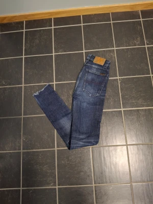 Nudie Jeans Skinny Blå W25 L32 - Säljer ett par blå skinny jeans från Nudie Jeans Co i storlek W25 L32. Jeansen har klassisk femficksdesign, slitna detaljer vid benslut och snygg tvätt med ljusare partier. Perfekta för dig som gillar en tajt passform och vill ha ett par jeans med edge.