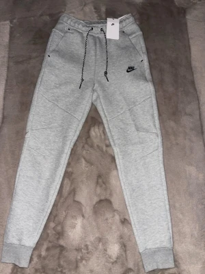 Grå Nike tech byxor  - Snygga grå mjukisbyxor från Nike med svart logga på benet och snörning i midjan. Byxorna har ribbade muddar vid bensluten, praktiska fickor och en svart dragkedja på sidan. Perfekta för chill eller träning, tillverkade i mjukt bomullsmaterial.