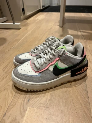 Air force 1 shadow - Nike Air Force 1 sneakers med grå mocka, vita partier i skinn och detaljer i neonrosa, grönt och svart. Swoosh-logga i svart med grön kant, samt AIR-tryck i rosa på hälen. Skorna har snörning och perforerad tå för extra ventilation.