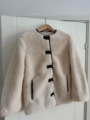 Beige teddyjacka från H&M - Säljer en beige teddyjacka från H&M med svarta detaljer och knappar längs framsidan. Jackan har rund hals, lång ärm och två fickor med svarta kanter. Materialet är mjukt och fluffigt, perfekt för kyliga dagar. Jackan har en loose passform och ger en cool vibe.