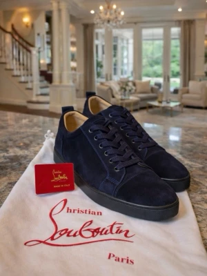 Mörkblå  Louboutins - Snygga mörkblå sneakers från Christian Louboutin i mjuk mocka med klassisk röd sula. Skorna har rund tå, svarta snören och diskret design som ändå sticker ut tack vare den ikoniska röda undersidan. Perfekta för dig som vill ha något exklusivt och stilrent.