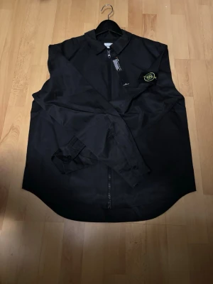 Svart overshirt från Stone Island - Säljer en svart overshirt från Stone Island med klassisk krage och dragkedja framtill. Jackan har den ikoniska Stone Island-loggan på ärmen och är tillverkad i ett lätt, syntetiskt material. Perfekt för lager-på-lager och stilrena outfits.