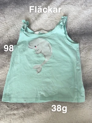 Mintgrön linnetopp med delfinmotiv - Söt mintgrön linnetopp med knyt på axlarna och ett glittrigt delfinmotiv framtill. Perfekt för varma dagar och enkel att matcha med shorts eller kjol. 🌸 Fläckar. Storlek 98