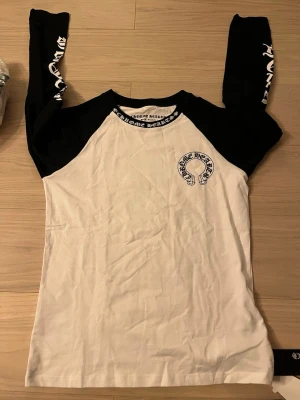 Chrome Hearts långärmad t-shirt svart/vit - Snygg långärmad t-shirt från Chrome Hearts i svart och vitt. Stor logga på bröstet och tryck längs ärmarna samt runt kragen. Klassisk baseball-modell med kontrastfärgade ärmar och mjukt bomullsmaterial. Perfekt för dig som gillar streetwear och vill sticka ut.