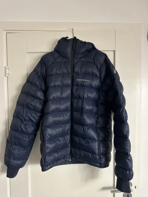 Mörkblå Peak Performance dunjacka - Säljer en mörkblå dunjacka från Peak Performance med huva och broderad logga på bröstet och ärmen. Jackan har quiltad design, dragkedja framtill och muddar vid ärmslut. Perfekt för kalla dagar 