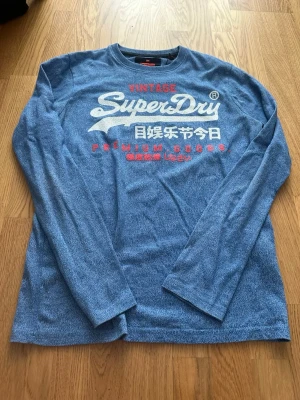 Blå tröja Superdry S - Säljer en blå långärmad tröja från Superdry med stort vitt och rött tryck framtill. Tröjan har rund halsringning och är gjord i mjuk bomullsmix. Perfekt för dig som gillar streetwear och vill ha en casual look.