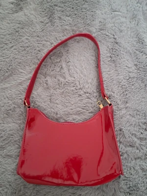 Röd axelväska i glansigt material - Säljer en snygg röd axelväska i glansigt syntetmaterial. Väskan har en enkel, stilren form med guldfärgade metalldetaljer och dragkedja upptill. Perfekt storlek för det viktigaste och ger en poppig touch till din outfit.