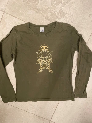 Olivgrön långärmad topp från Stüssy - Olivgrön långärmad topp från Stüssy i bomull med gult tryck. Vintage! Liten i storleken. 