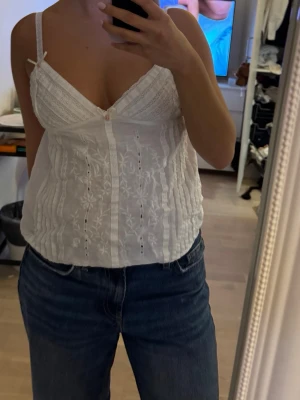Vit broderad linnetopp med spets - Säljer en söt vit linnetopp med tunna axelband och  rosetter. Toppen har broderade blommor framtill, spetsdetaljer vid bysten och en v-ringad skärning. 