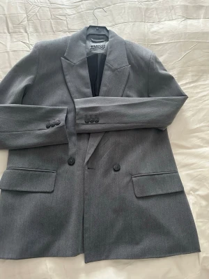 Grå kavaj från Amisu   - Snygg grå kavaj med klassisk krage och dubbelknäppning framtill. Kavajen har två fickor med lock och är helfodrad. Perfekt för dig som vill ha en stilren och tidlös look. Passar till både jeans och kostymbyxor.